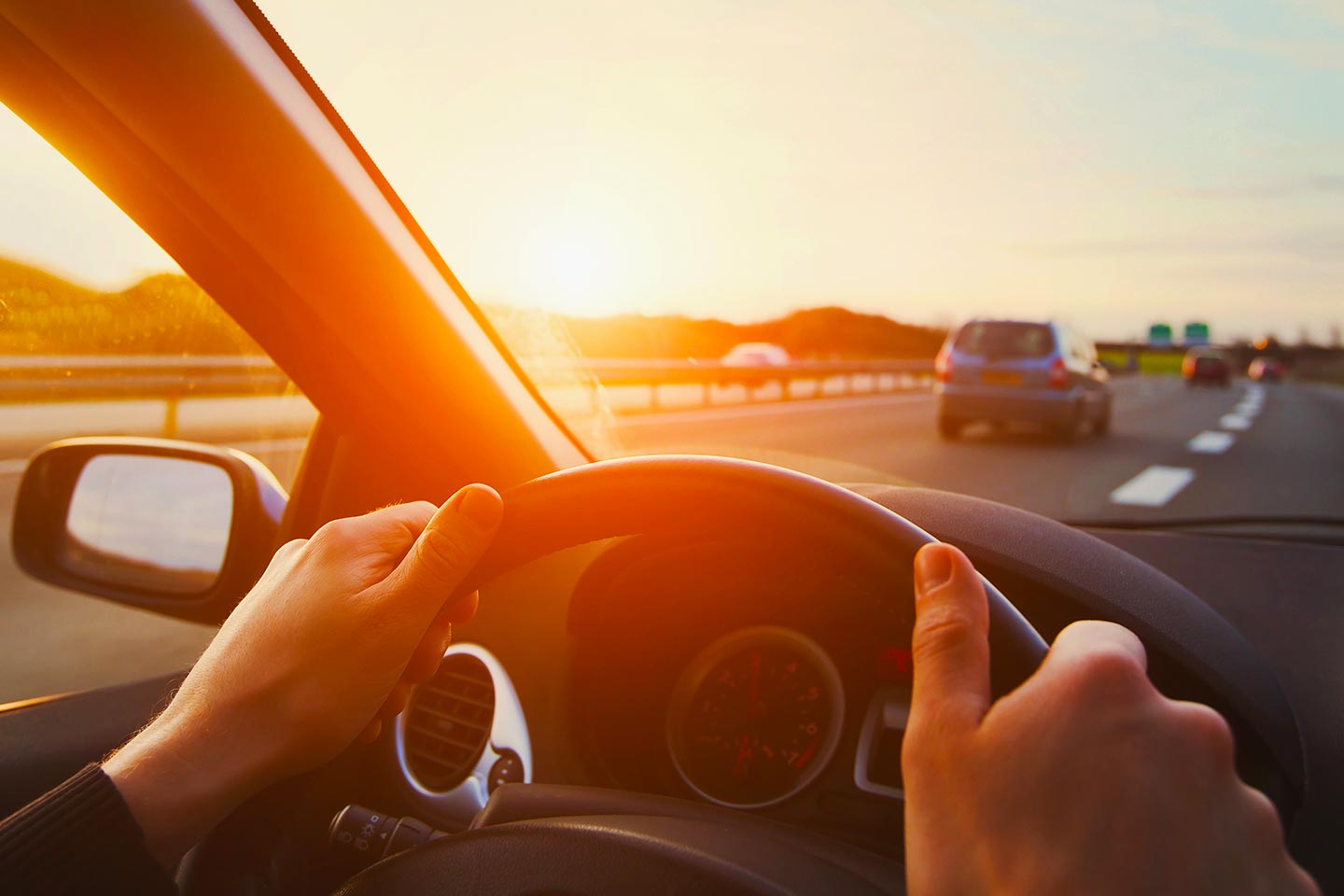 A person's hands on the steering wheel driving into the sunset.