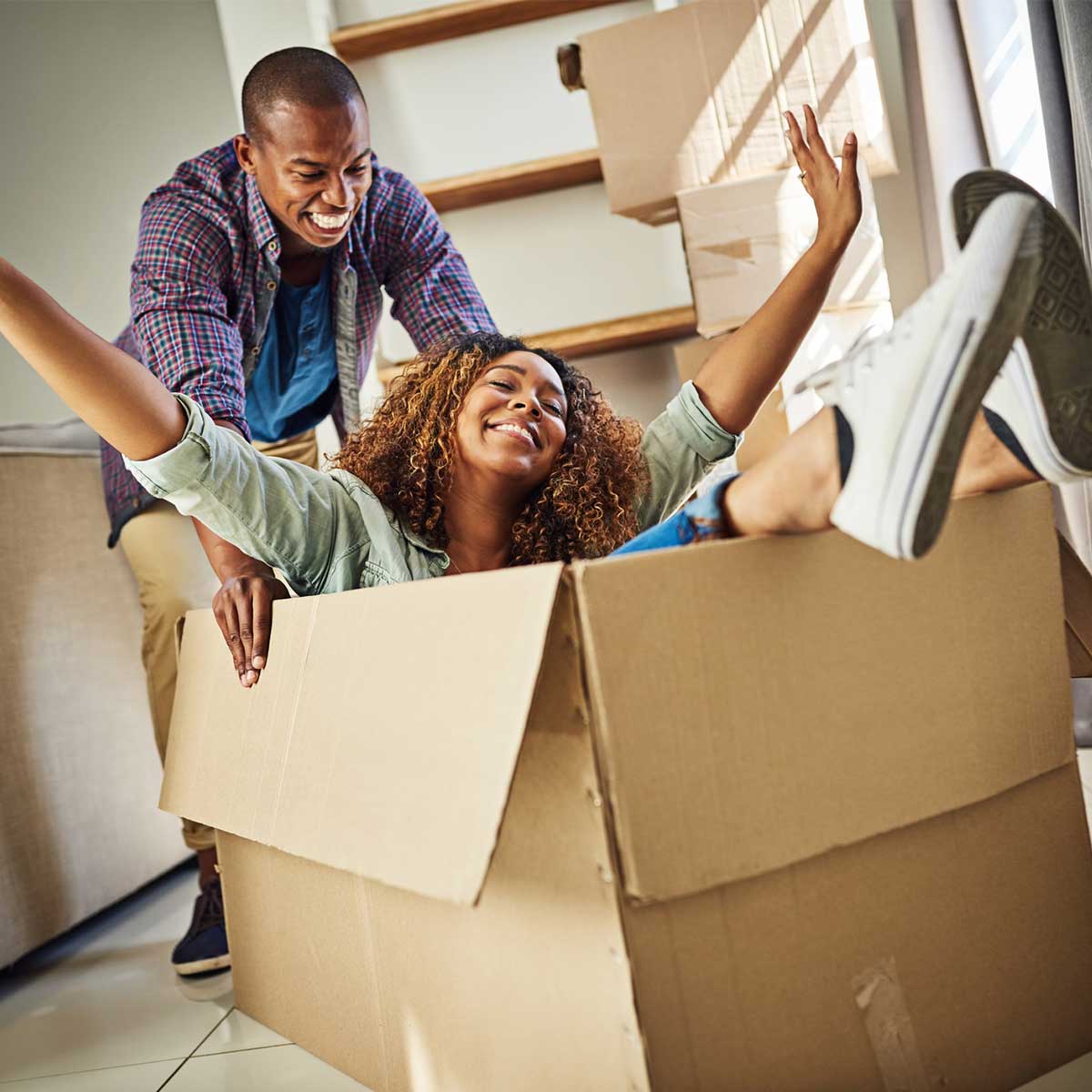 Cheerful young woman inside of a box with her partner pushing the box inside an apartment